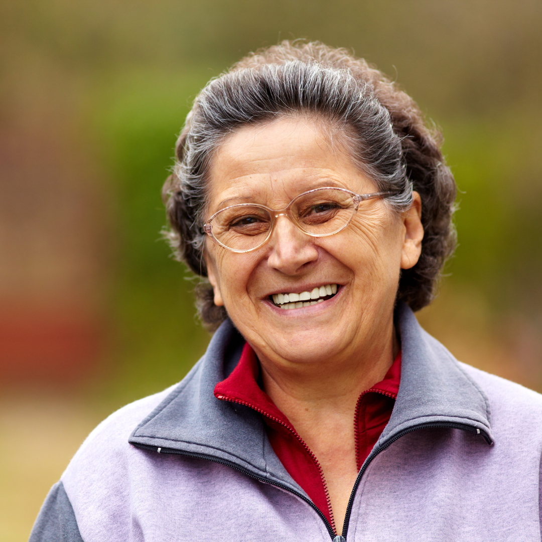 Smiling elderly woman with glasses, wearing a gray jacket with red collar, outdoors with blurred fall foliage background.