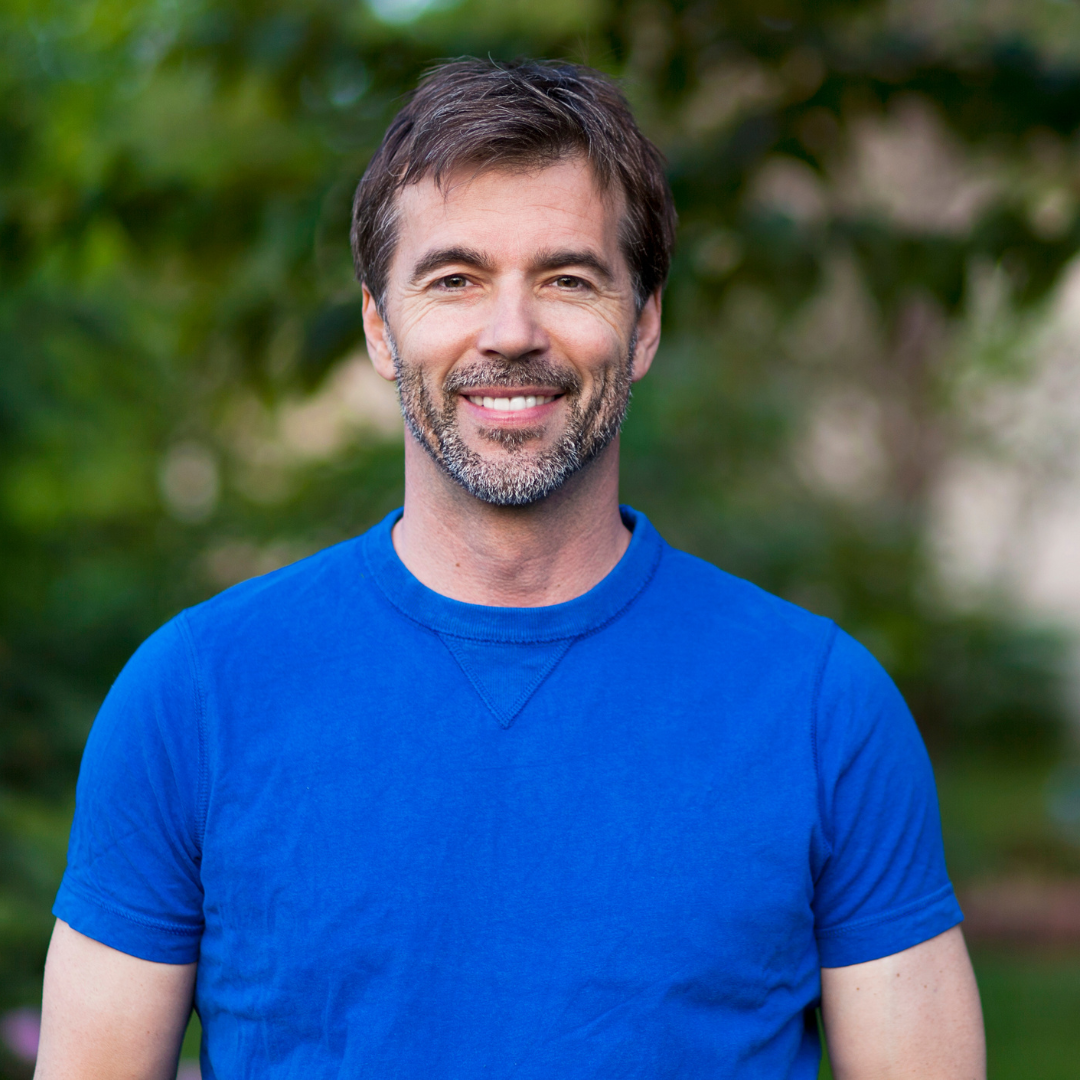 A smiling man with brown hair and a beard, wearing a blue t-shirt, standing outdoors with blurred green foliage in the background.