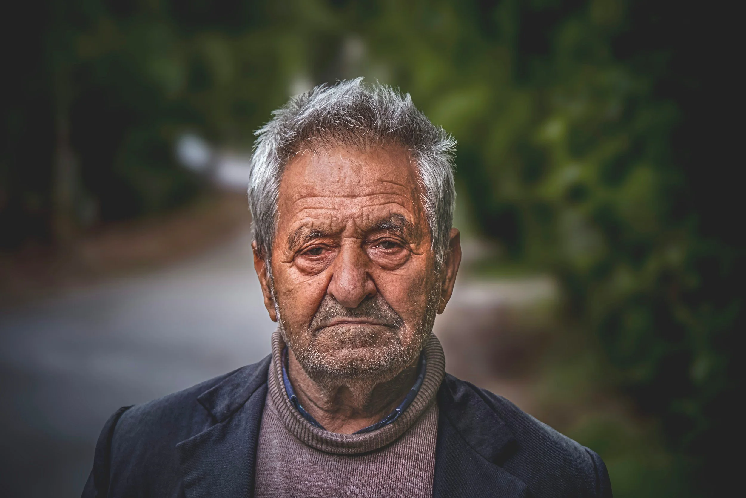 Close-up of an elderly man with gray hair and a serious expression, outdoors with blurred green foliage in the background.