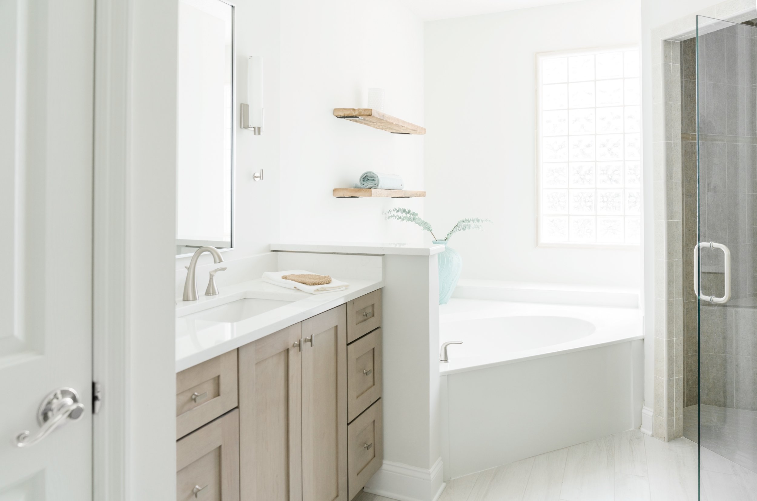 Bright bathroom with a vanity and faucet, a bathtub with a window behind it, and a glass shower door.