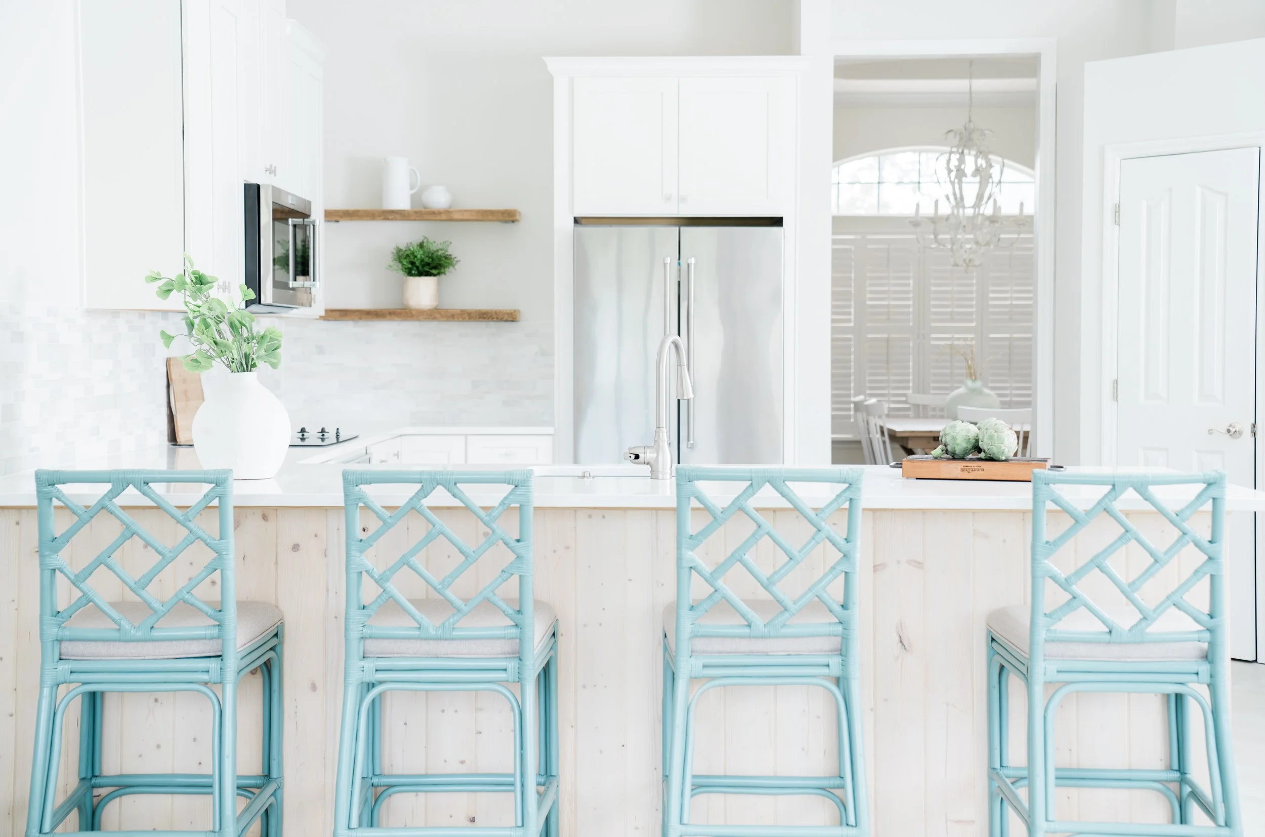 White kitchen with light blue wicker barstools, white countertops, stainless steel refrigerator, and open shelving with decorative items.