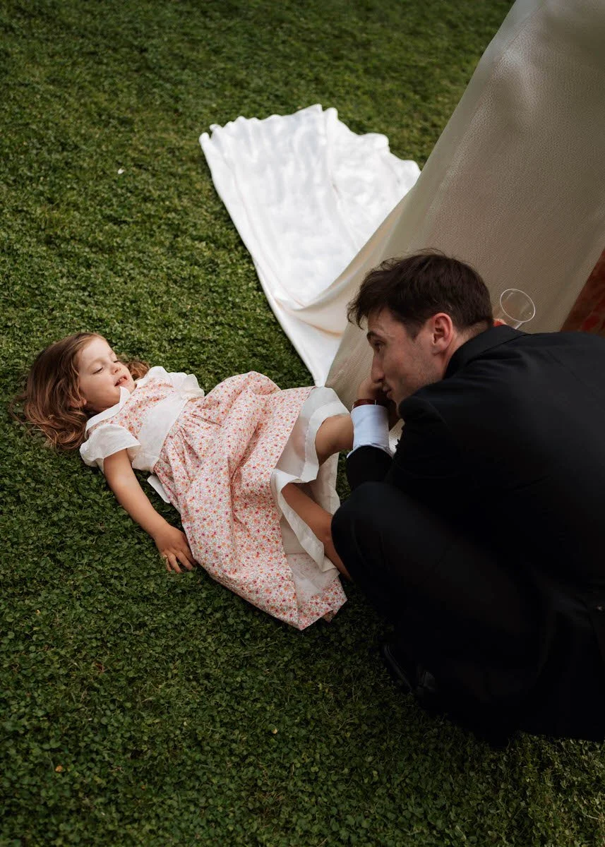 A man in a black suit kneeling next to a young girl lying on the grass. The girl has red hair and is wearing a white and pink floral dress. The man appears to be praying or speaking softly to the girl, who is resting with her eyes closed.