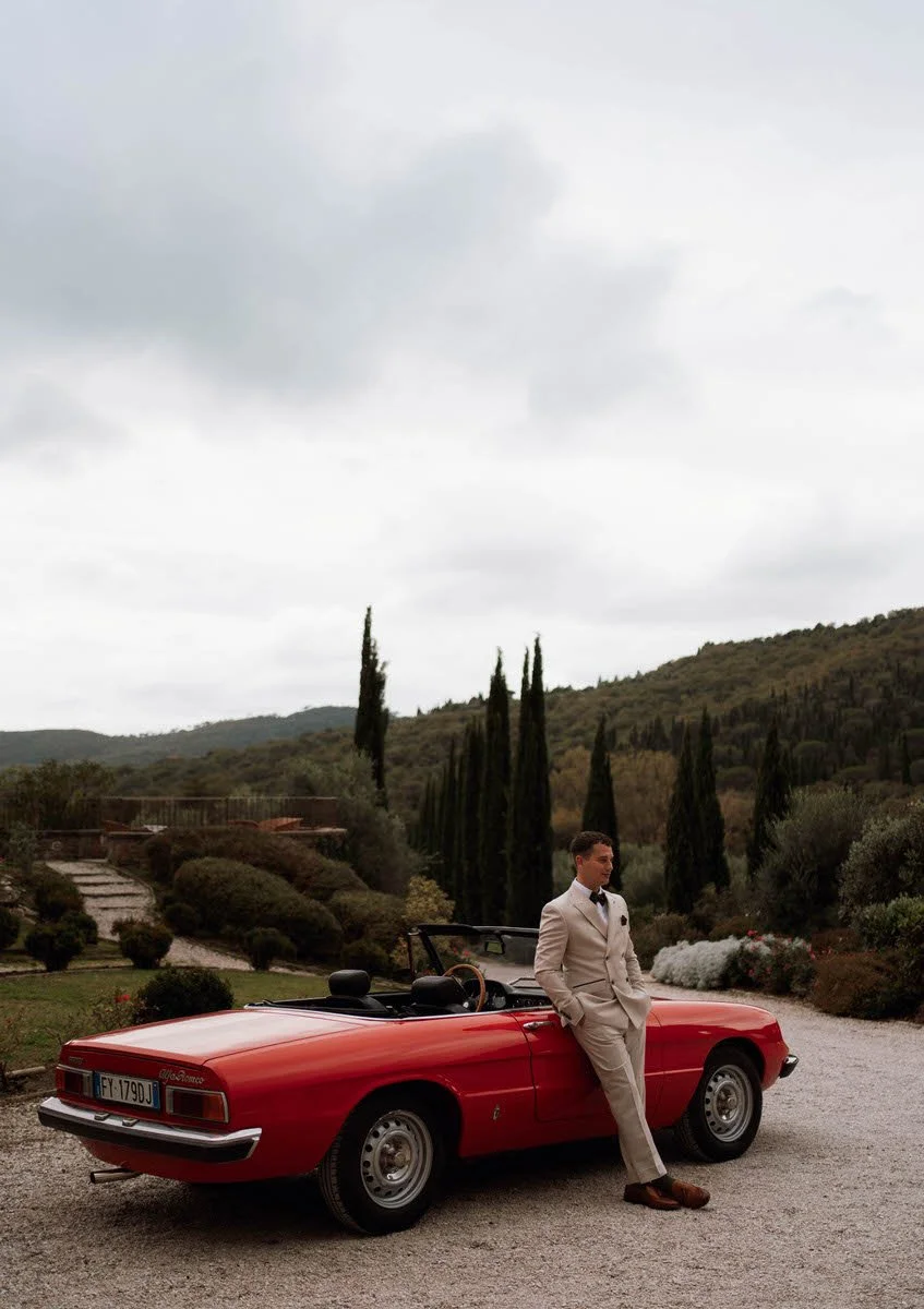 A man in a beige suit with a bowtie standing next to a red convertible car in a scenic outdoor area with trees and hills under a cloudy sky.