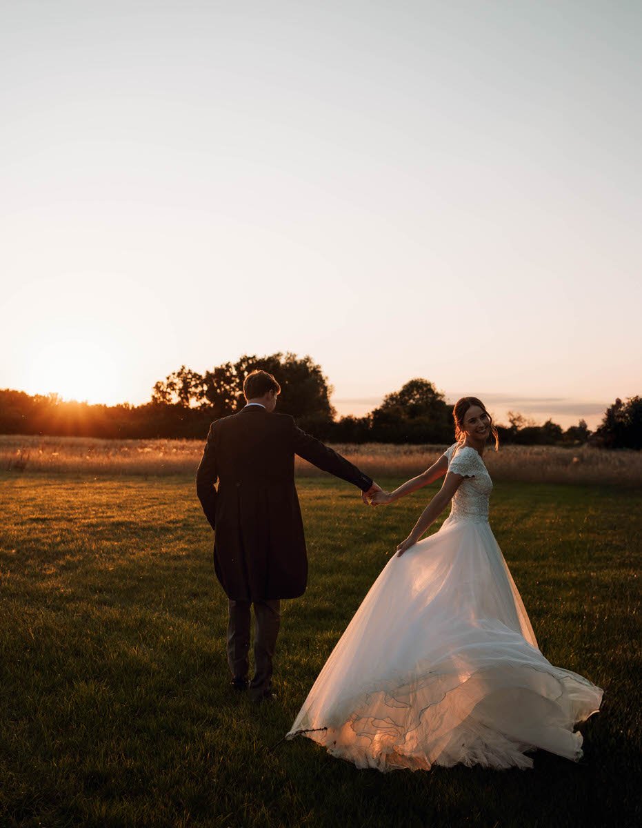 A bride and groom holding hands and walking in a field during sunset, with the bride smiling and wearing a white wedding dress.