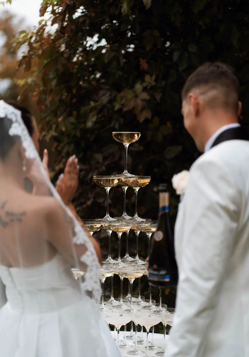 A bride and groom dressed in wedding attire watching a champagne tower at their wedding celebration outdoors with dark foliage in the background.
