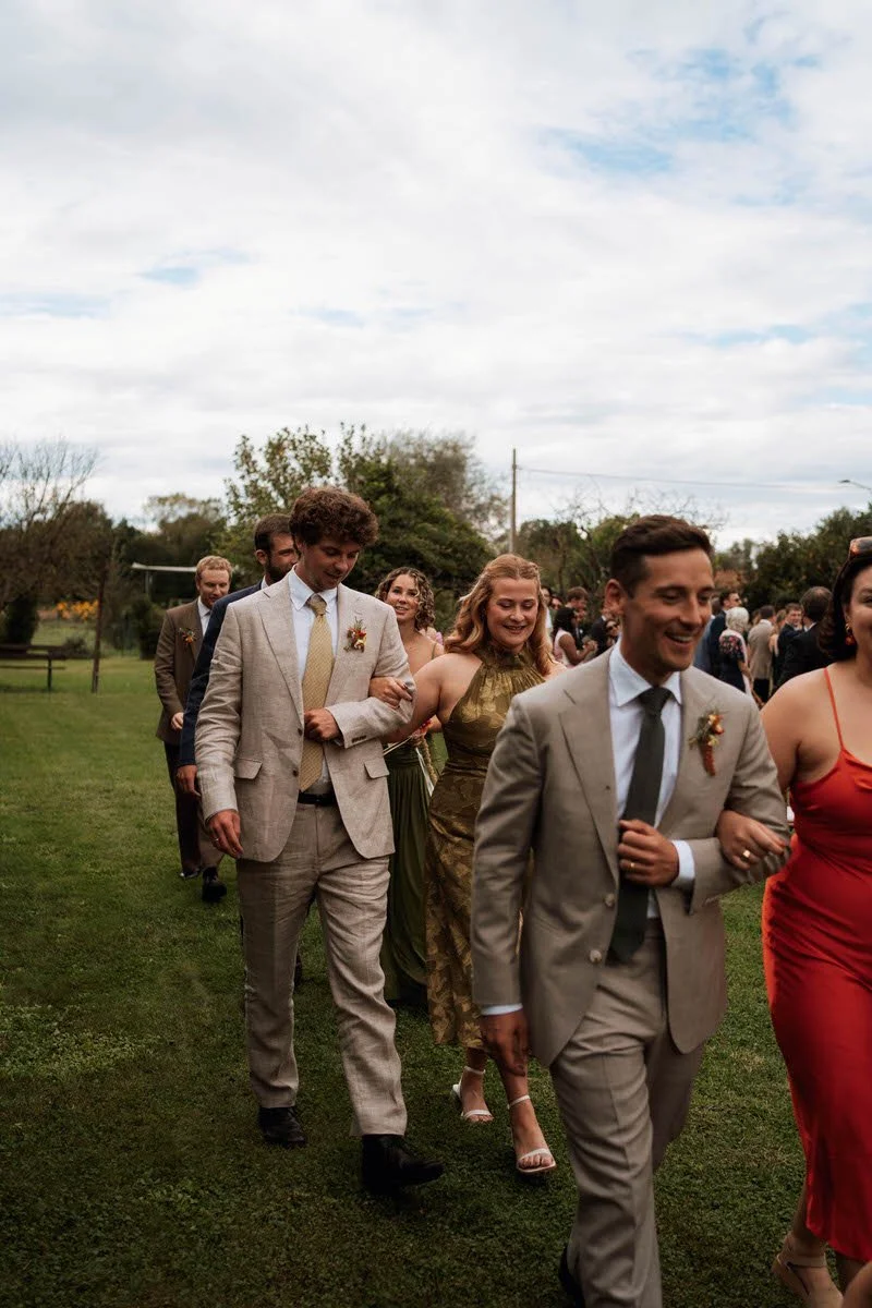 Group of people dressed in formal attire walking outdoors on a grassy field, likely at a wedding or special event under a cloudy sky.