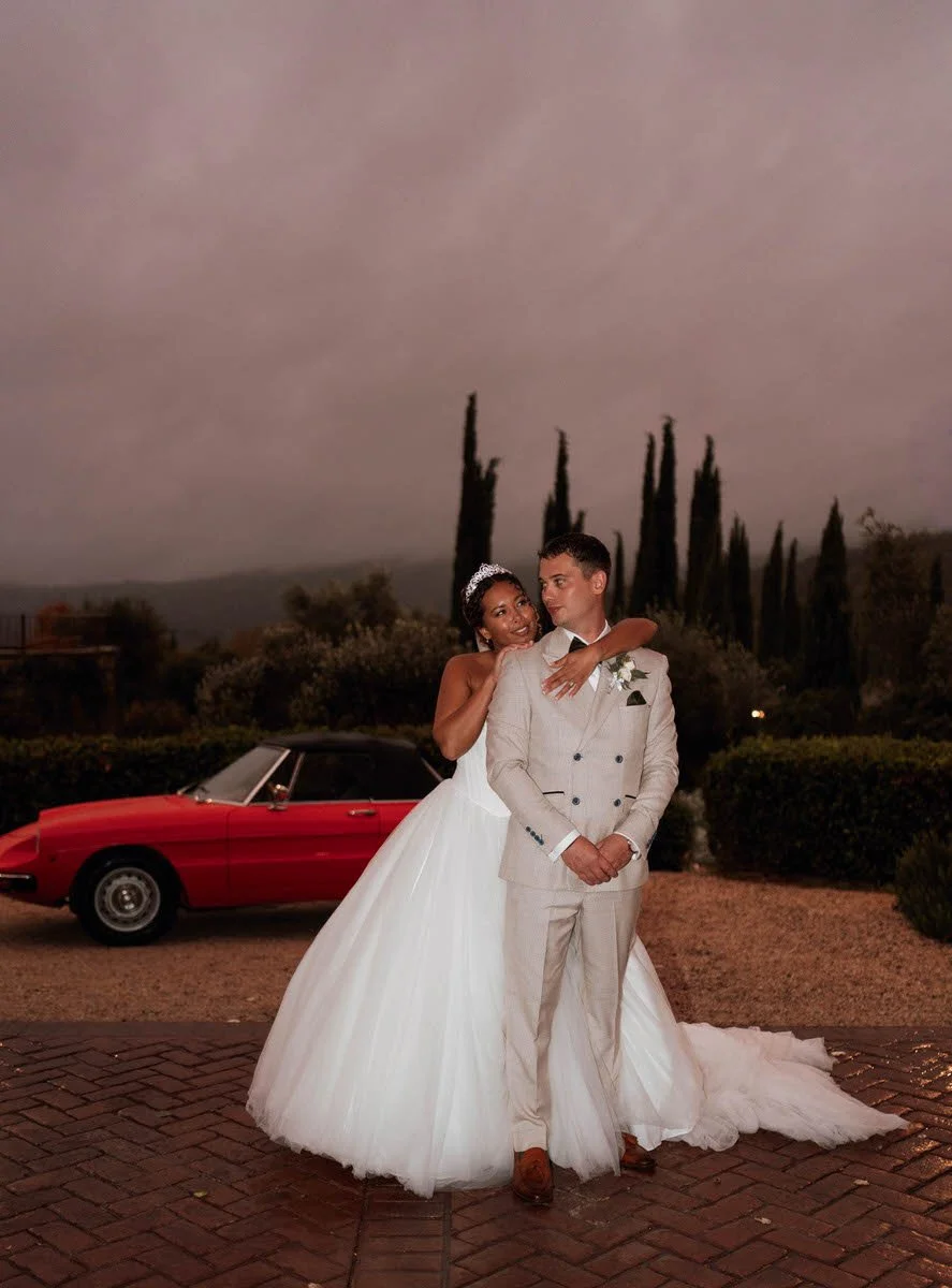 A bride and groom standing outdoors during dusk. The bride is in a white wedding gown with a tiara, hugging the groom, who is dressed in a light-colored suit. In the background, there are tall trees, bushes, and a red vintage convertible car on a bri