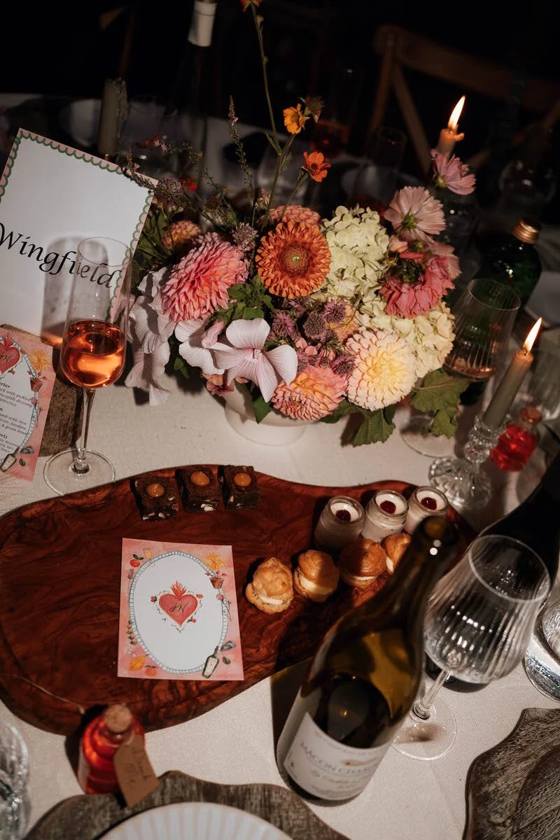 A table setting with a large floral centerpiece featuring pink, orange, and white flowers, lit candles, wine bottles, glasses, and assorted desserts on a wooden platter, with decorative cards and tableware.