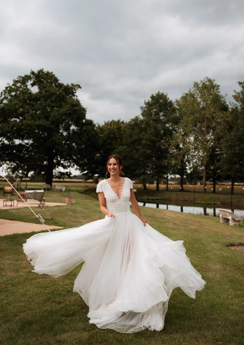A smiling woman in a white wedding dress twirling outdoors on a grassy area with trees and a pond in the background under a cloudy sky.