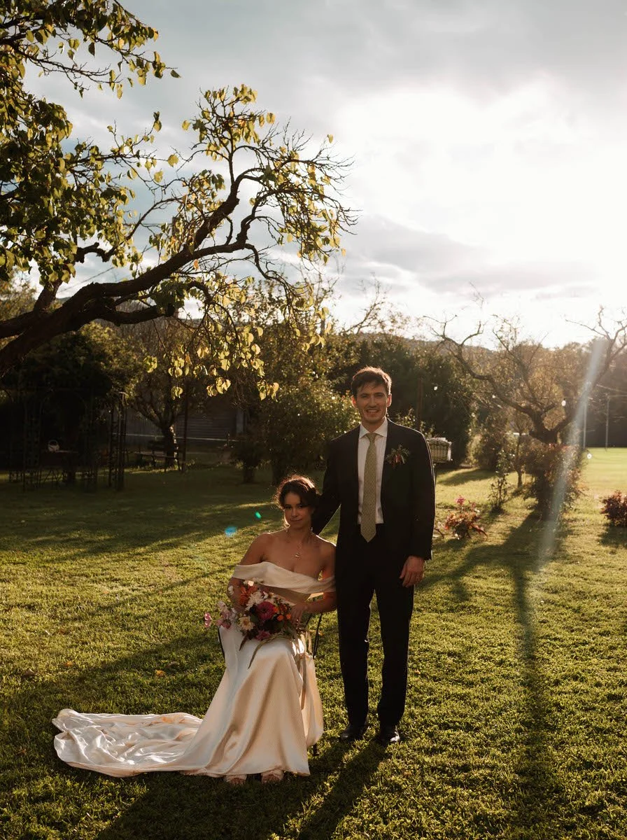 A bride in a white wedding dress sitting on a chair holding a bouquet of flowers, and a groom in a suit standing beside her, outdoors in a grassy area with trees and a bright sky.