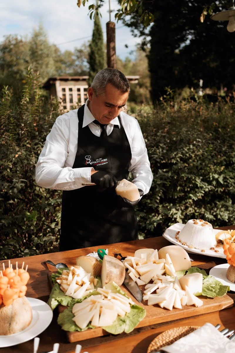 Man slicing cheese outdoors at a table with cheese, bread, and other appetizers.