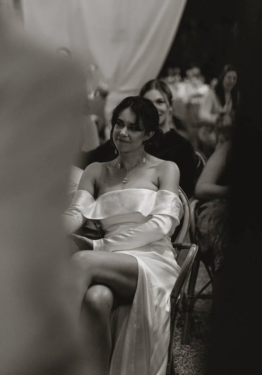 A woman wearing an off-the-shoulder satin dress sitting at a formal gathering or event with other guests in the background. The photo is in black and white.