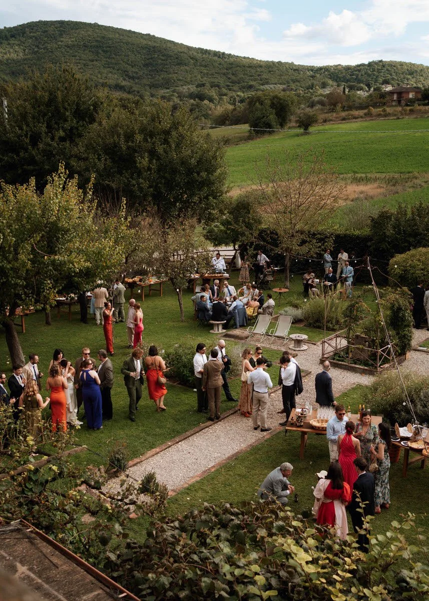 Outdoor wedding reception or party in a garden surrounded by trees and hills, with guests socializing, eating, and enjoying drinks.