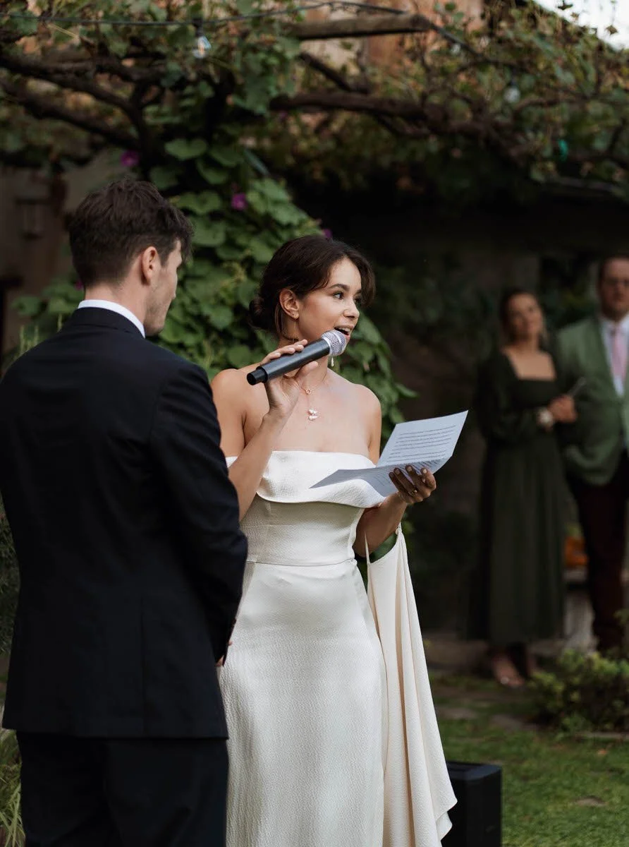 A bride giving a speech at her outdoor wedding ceremony, holding a microphone and reading from a paper, standing beside the groom in a tuxedo with barefoot guests in the background.