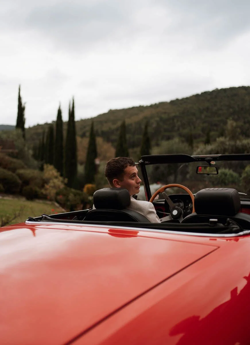 A young man sits in the driver's seat of a red convertible car, looking over his shoulder with a scenic view of trees, hills, and an overcast sky in the background.