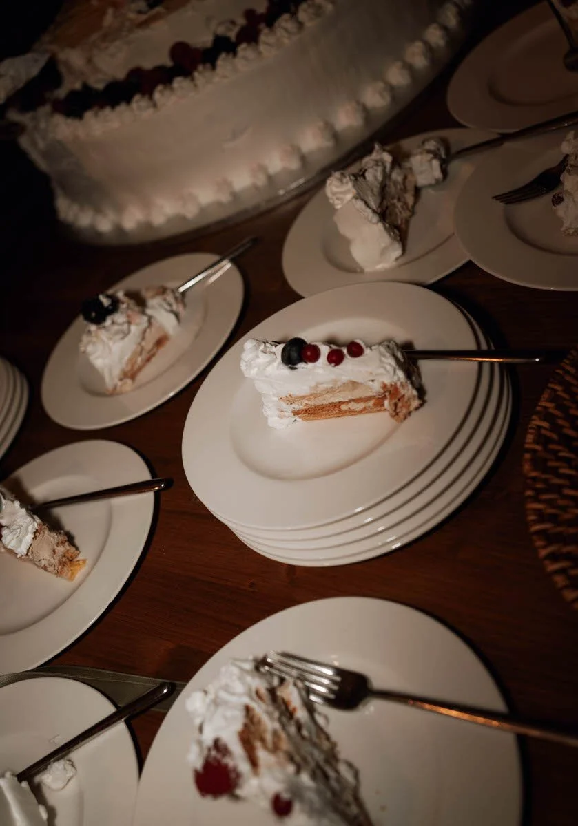 Several slices of birthday cake with whipped cream and berries on white plates, with a large decorated cake in the background.