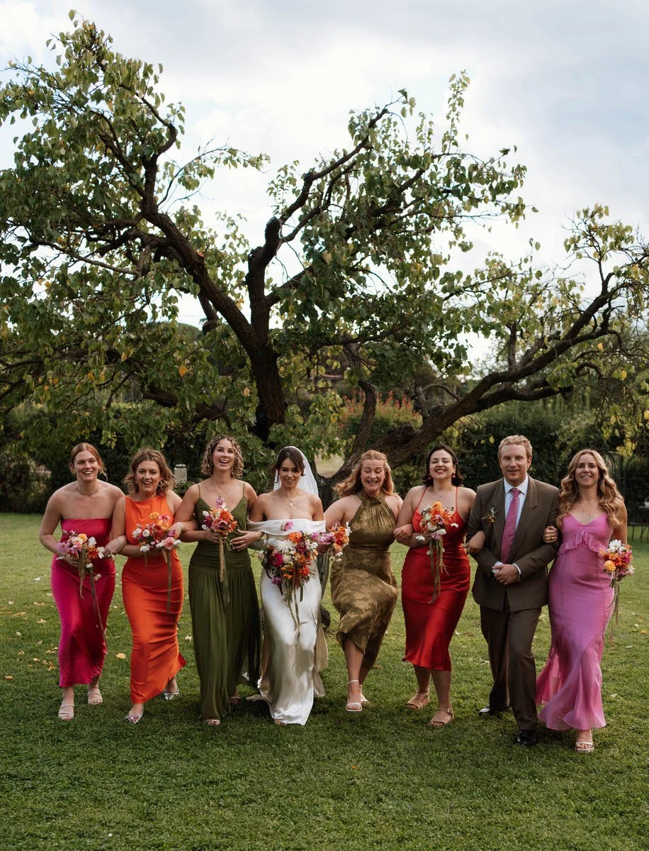 Group of women in colorful dresses and a man in a suit walking arm-in-arm outdoors near a large tree during a celebration or wedding.