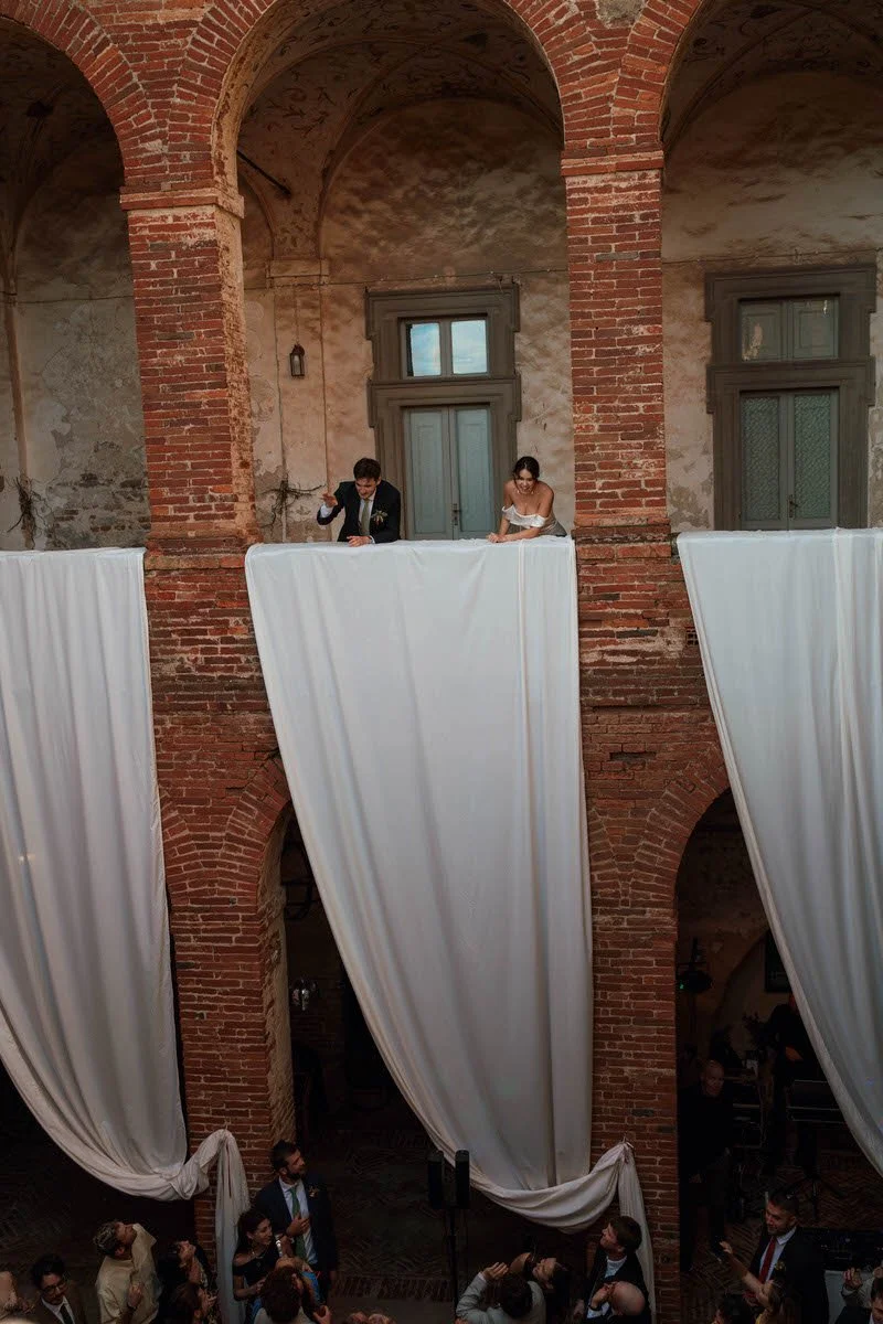 A man and woman, possibly at a wedding or celebration, standing behind a white draped table on a balcony in a rustic brick building with arched openings, while guests are gathered below.