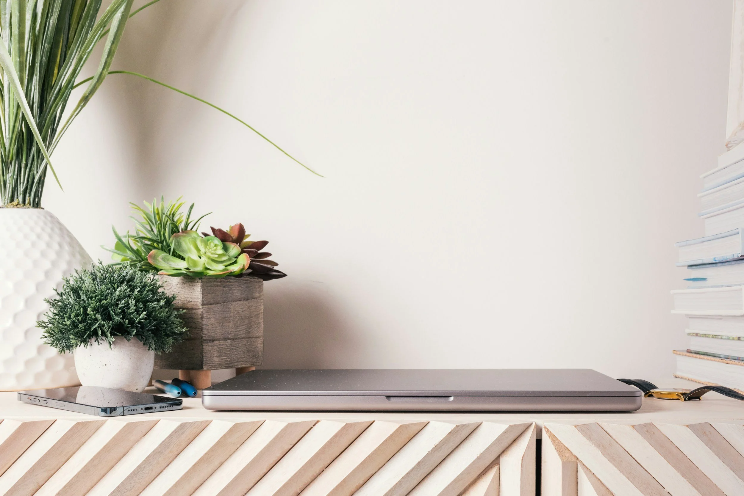 Desk with closed silver laptop, potted plants, and a stack of books against a plain white wall.