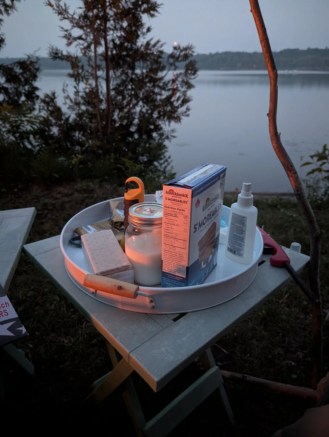 A firelit tray of s'mores supplies in front of a lake at dusk: graham crackers, chocolate, marshmallows, milk in mason jars, and - crucially - bug spray.