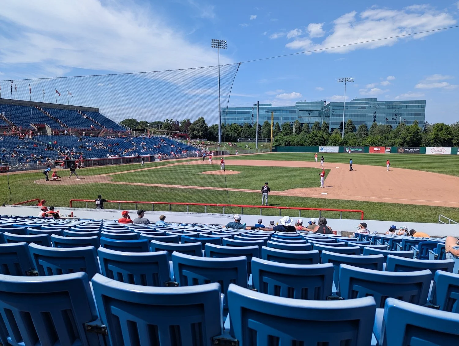 Blue plastic ball park seats in the foreground, and beyond a summer Frontier League baseball game with the home team in red-and-white in the field.