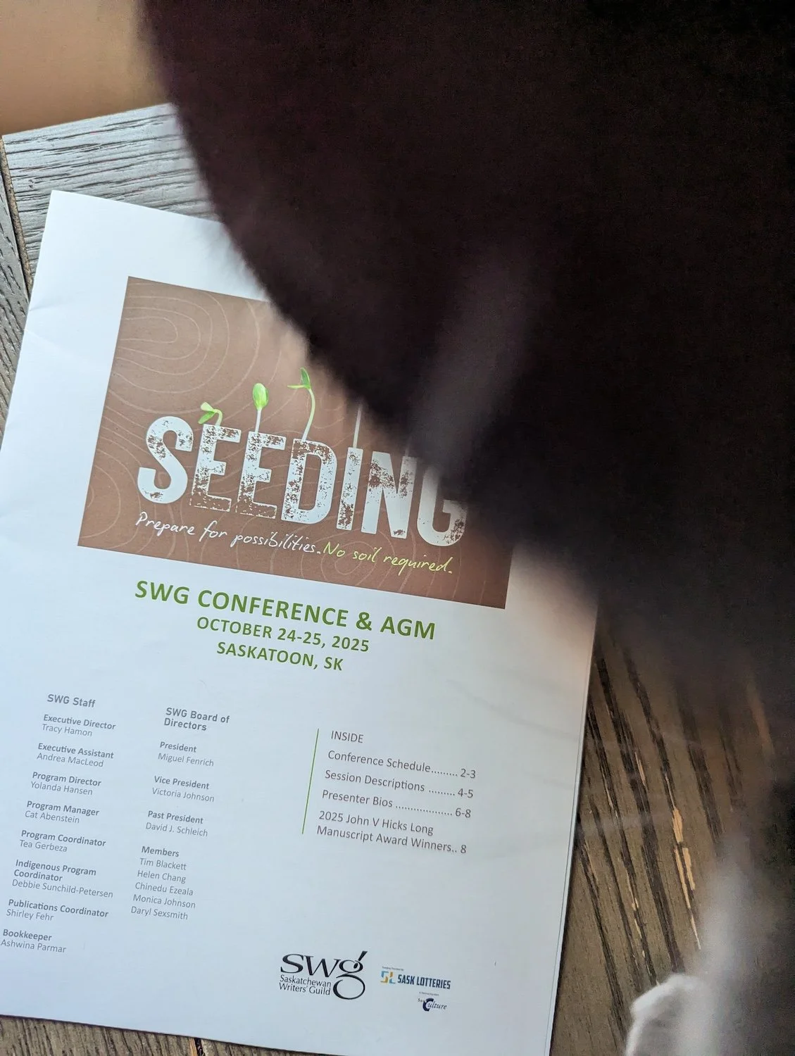 A photo of the 2025 SWG AGM and Conference program, photobombed by two cats whose blurry sides, whiskers and paw are visible - one is black and the other is grey.