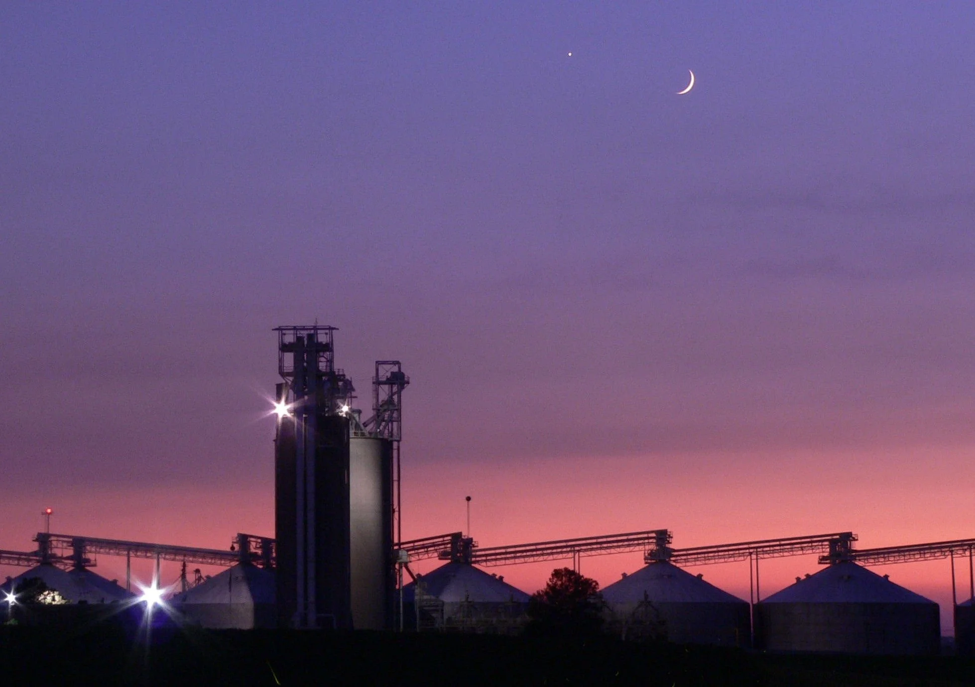 Venus and the Moon in a purple dusk sky above a pink sunset skyline, with grain elevators and silos in the foreground.