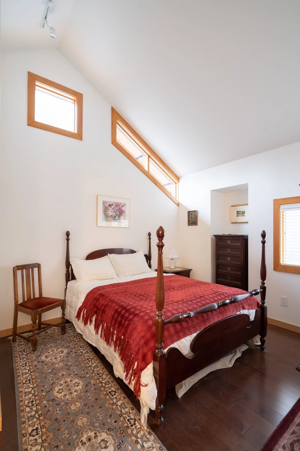 A low four-poster bed in a white bedroom with lace and other old-fashioned textiles on the dark wood furniture.