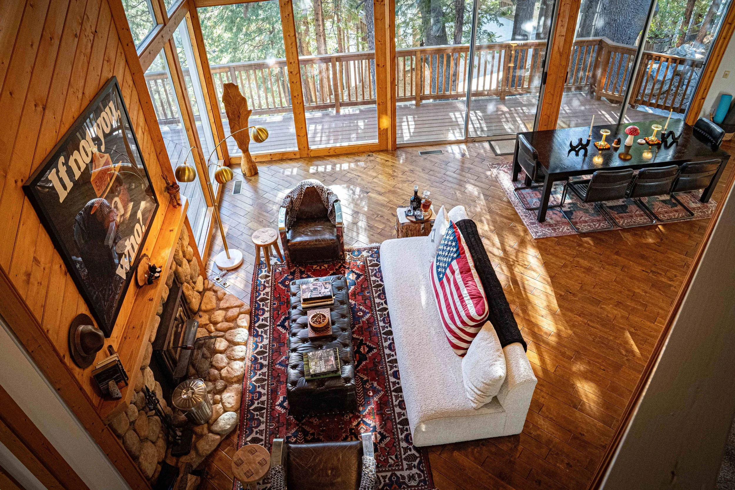 Living room with wooden walls, a stone fireplace, and large glass windows overlooking a deck and trees. Contains a black leather armchair, a white sofa with an American flag blanket, a patterned rug, a wooden coffee table with magazines, a small side