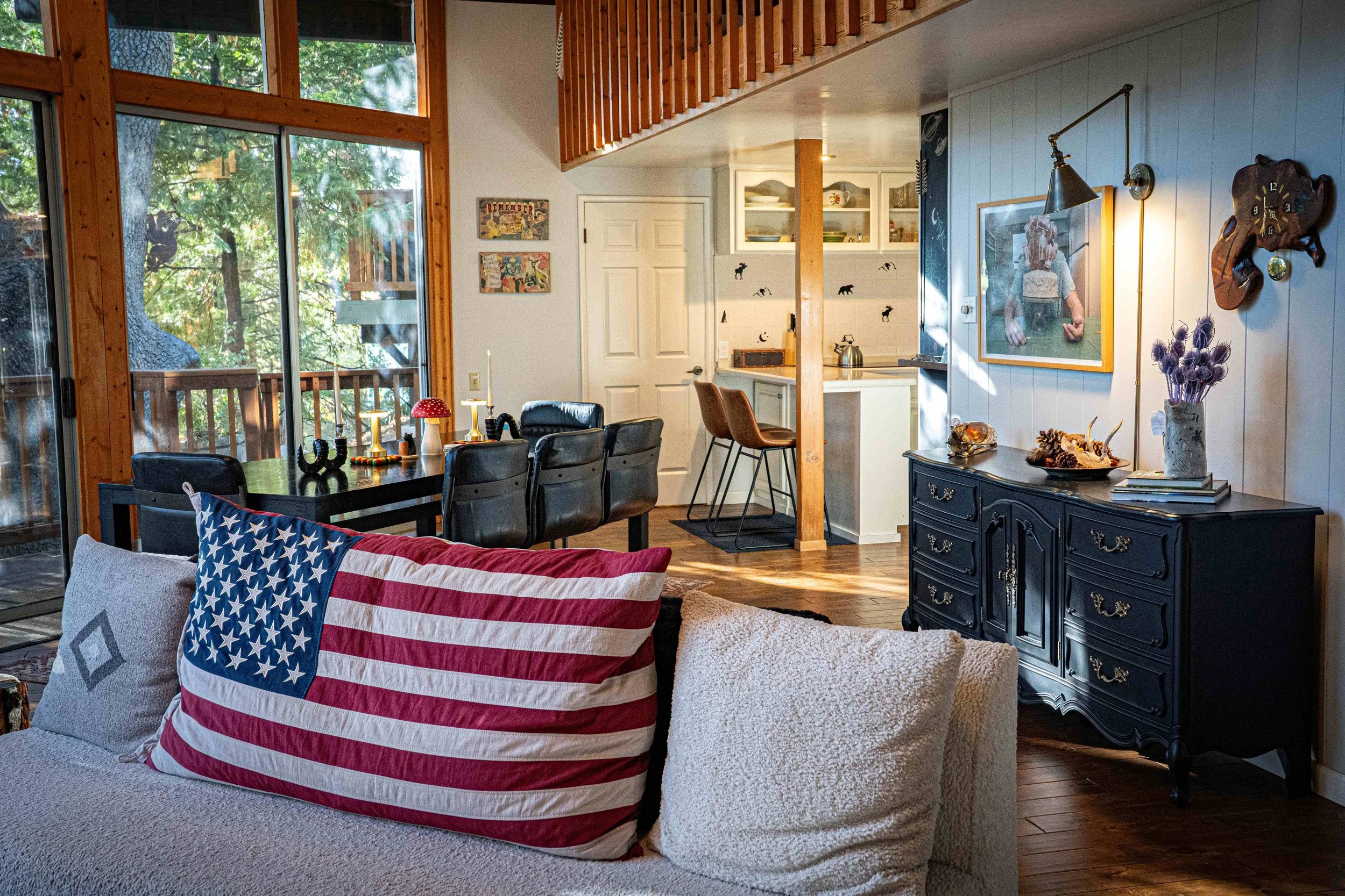Living room with large windows showing trees outside, a black dining table with chairs, a couch with an American flag pillow, a black sideboard decorated with flowers and shells, and a wall with artwork and a framed photo, adjacent to a kitchen area 