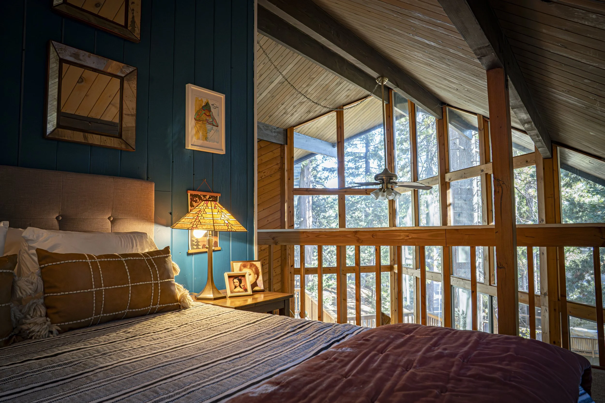 A cozy loft bedroom with a bed decorated with pillows, a wooden nightstand with a lamp and framed photos, wooden walls and ceiling, large windows showing trees outside, and a ceiling fan.
