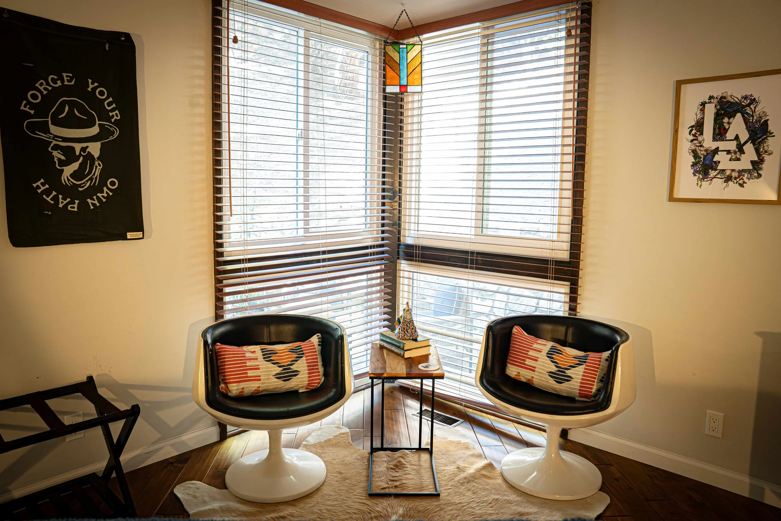 Cozy corner with two modern black and white swivel chairs with patterned pillows, a wooden side table with books and a decorative object, large windows with wooden blinds, a colorful hanging lamp, wall art including a black banner with a skull and ha