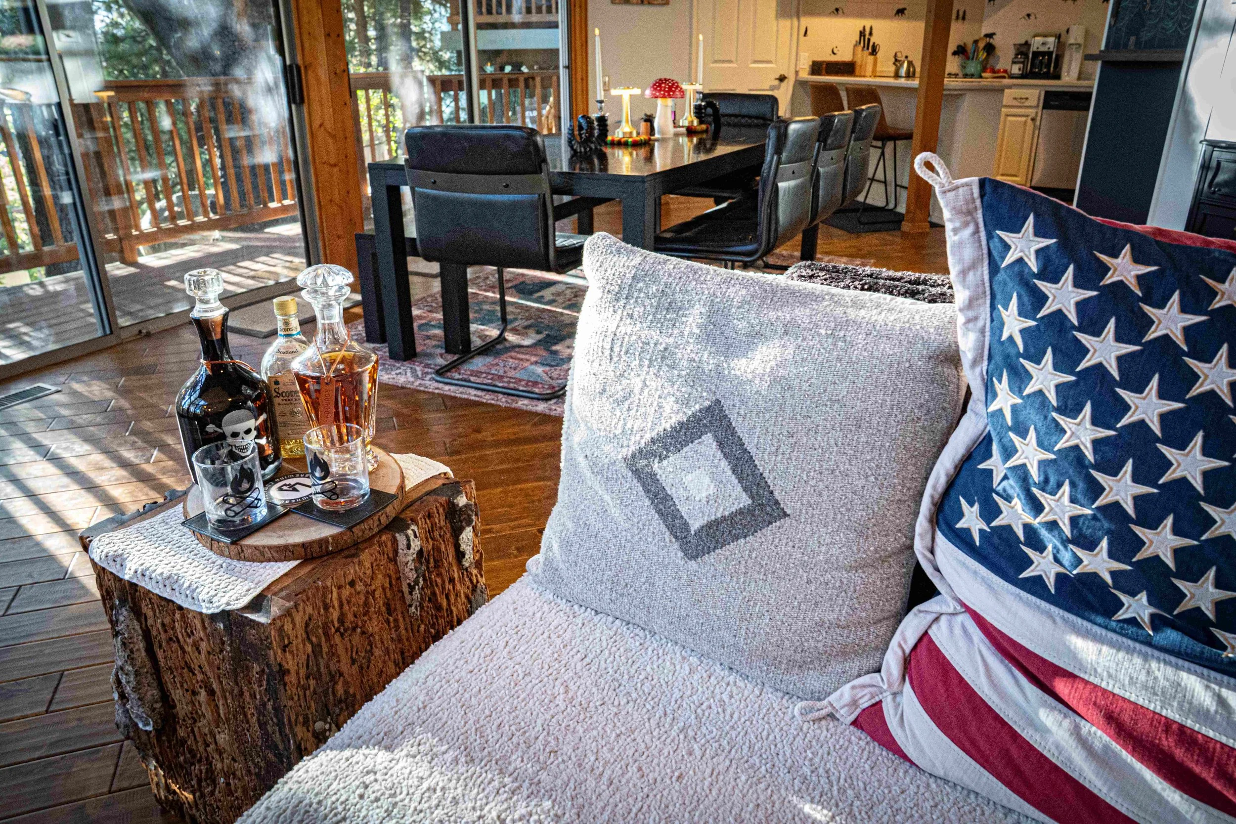 Indoor living space with wooden flooring, a collection of liquor bottles and glasses on a tree trunk side table, a beige sofa with patriotic pillows, a dark dining table with black chairs, and a sliding glass door leading to a wooden deck outside.