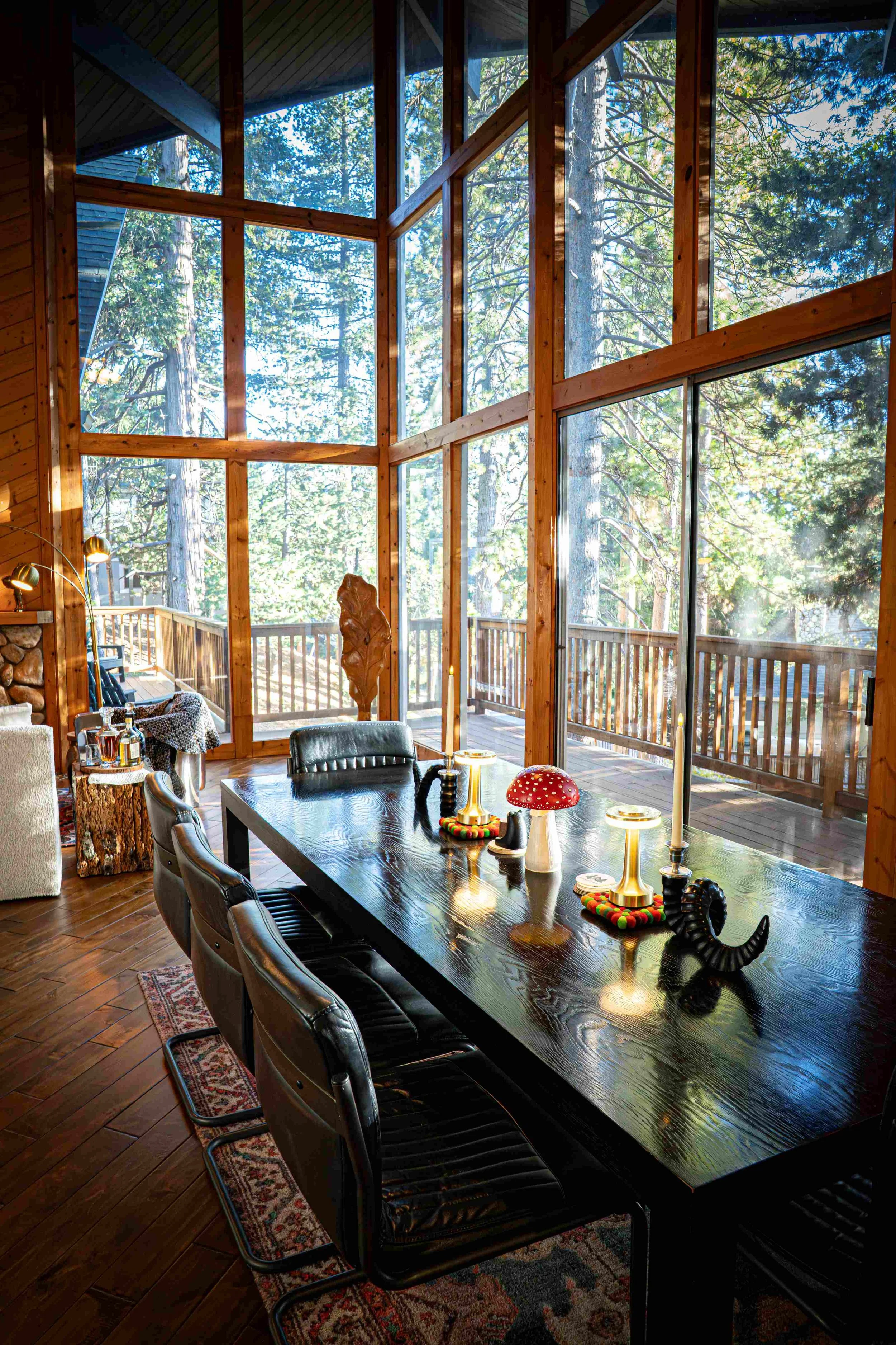 Dining room with a large wooden table, black chairs, and decorative items including lamps, candles, and figurines. Floor-to-ceiling windows reveal a forest outside.