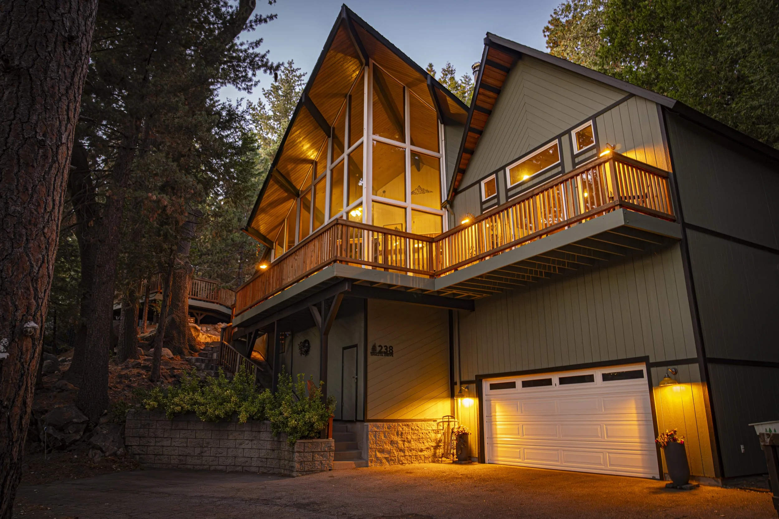 A modern multi-story house A-frame illuminated with warm lights, surrounded by trees, with a garage and a balcony, at dusk.