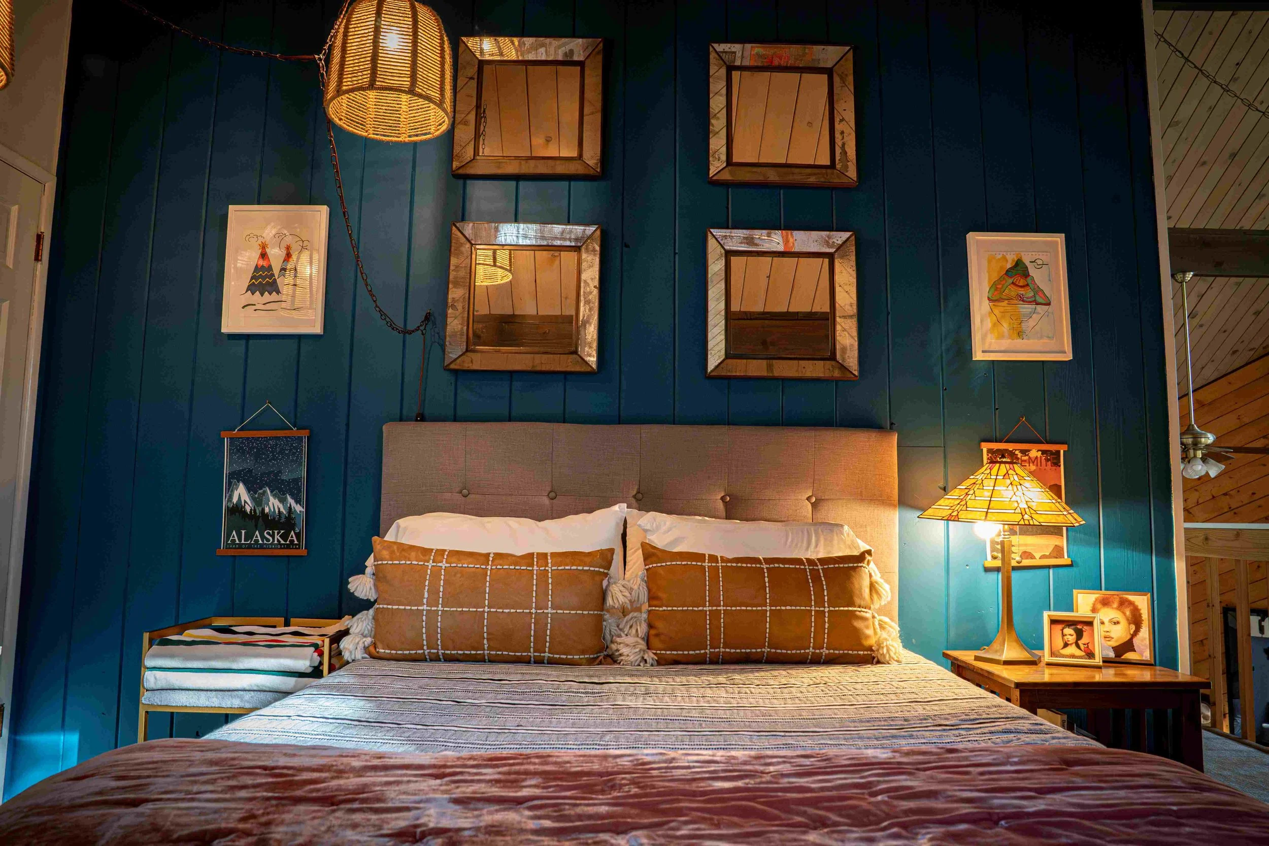 A cozy bedroom with a blue paneled wall, four square mirrors arranged in a grid, two framed mountain-themed artworks, a lamp with a stained glass shade, and a bedside table with framed photographs. The bed has a tan headboard and brown plaid pillows.