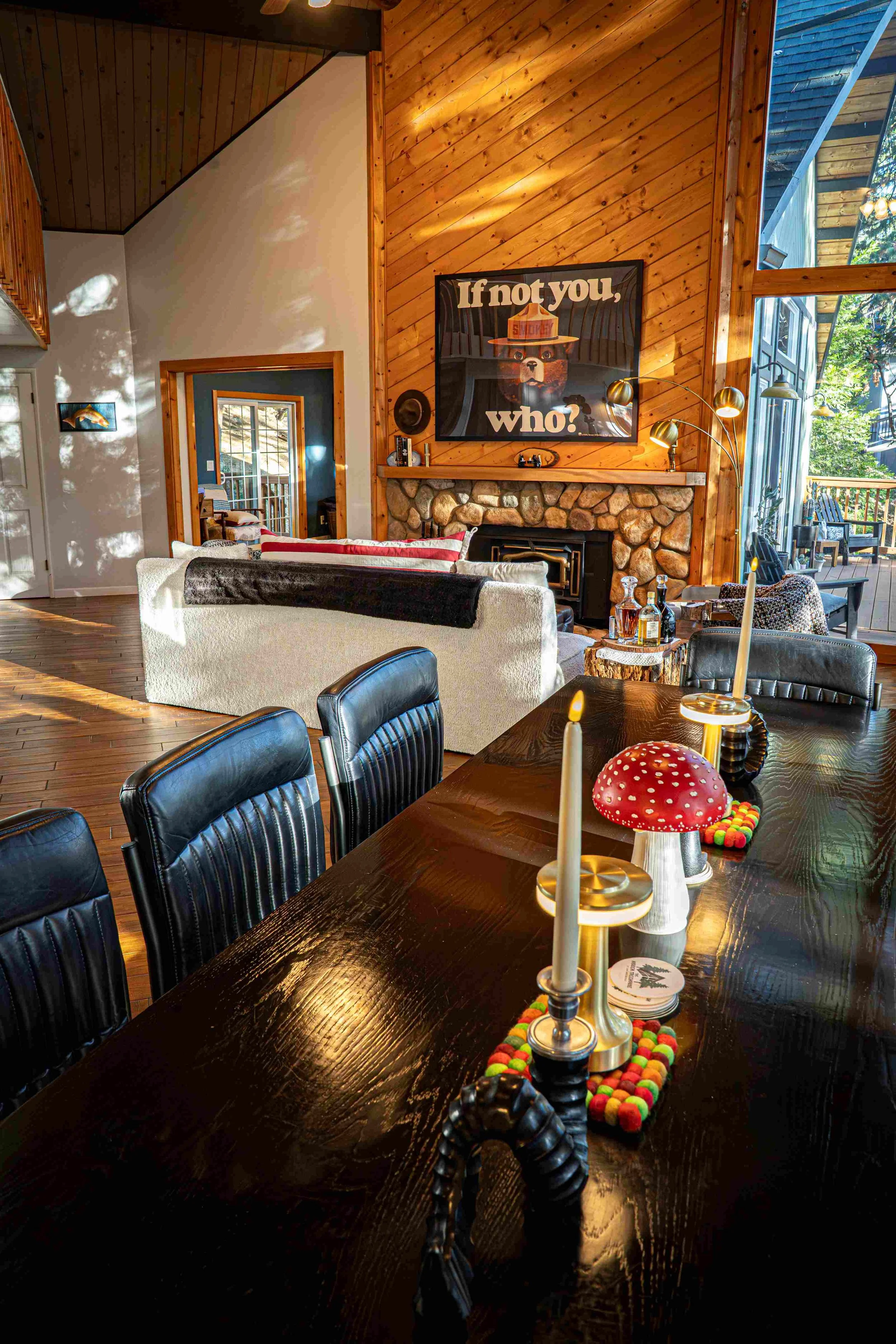 The image shows a cozy living room area with a wood-paneled wall, a stone fireplace, and large windows allowing natural light. There is a white couch with red and black pillows facing the fireplace, and a dining table in the foreground with candles, 