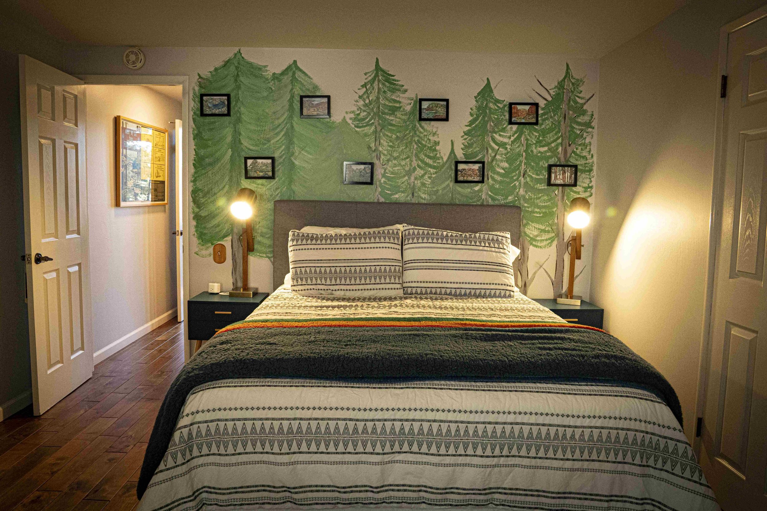 Bedroom with a mural of green trees on the wall behind the bed. The bed has patterned pillows and a multicolored blanket. There are nightstands with lamps on either side of the bed, and framed pictures above the mural.