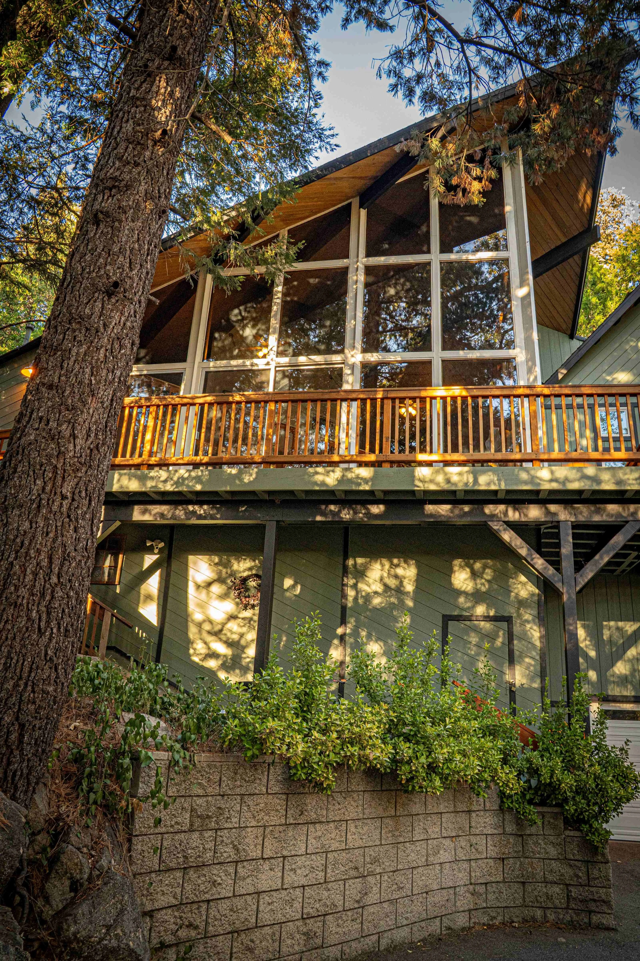 A modern house with a large glass front, wooden balcony, and green exterior, surrounded by tall trees and bushes.