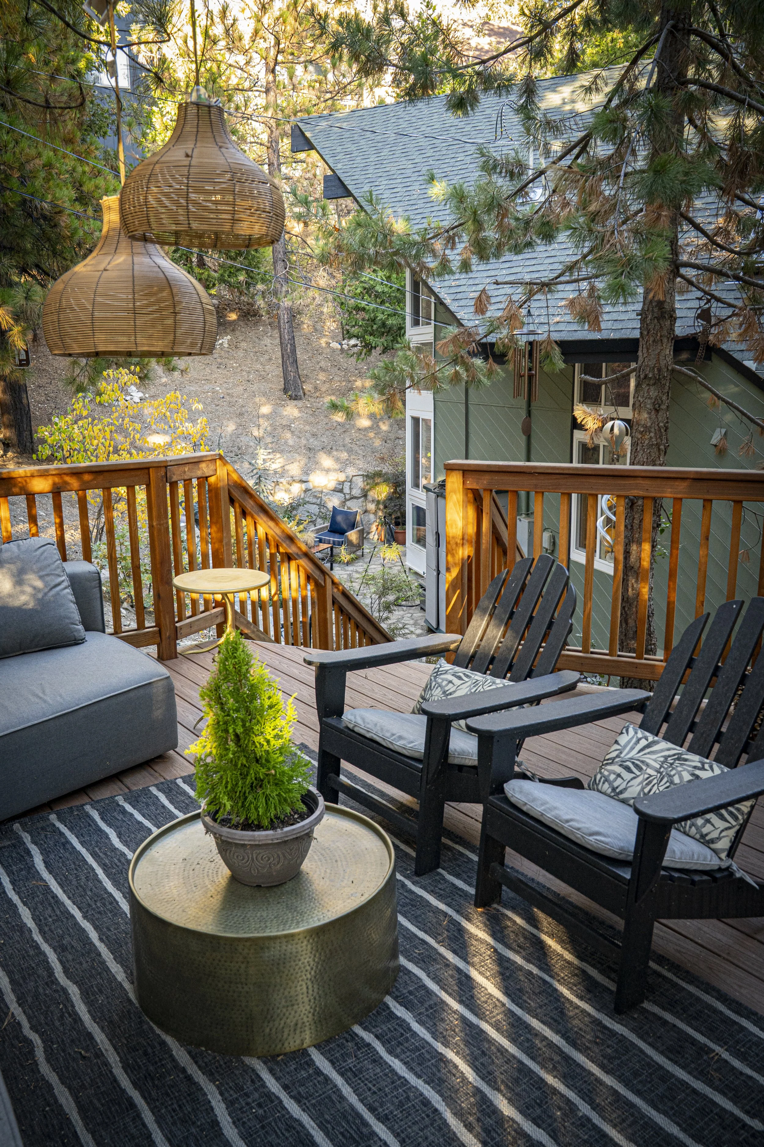 An outdoor deck with black Adirondack chairs, a gray cushioned sofa, a small round side table, and a large potted plant, with a view of trees and a house in the background.