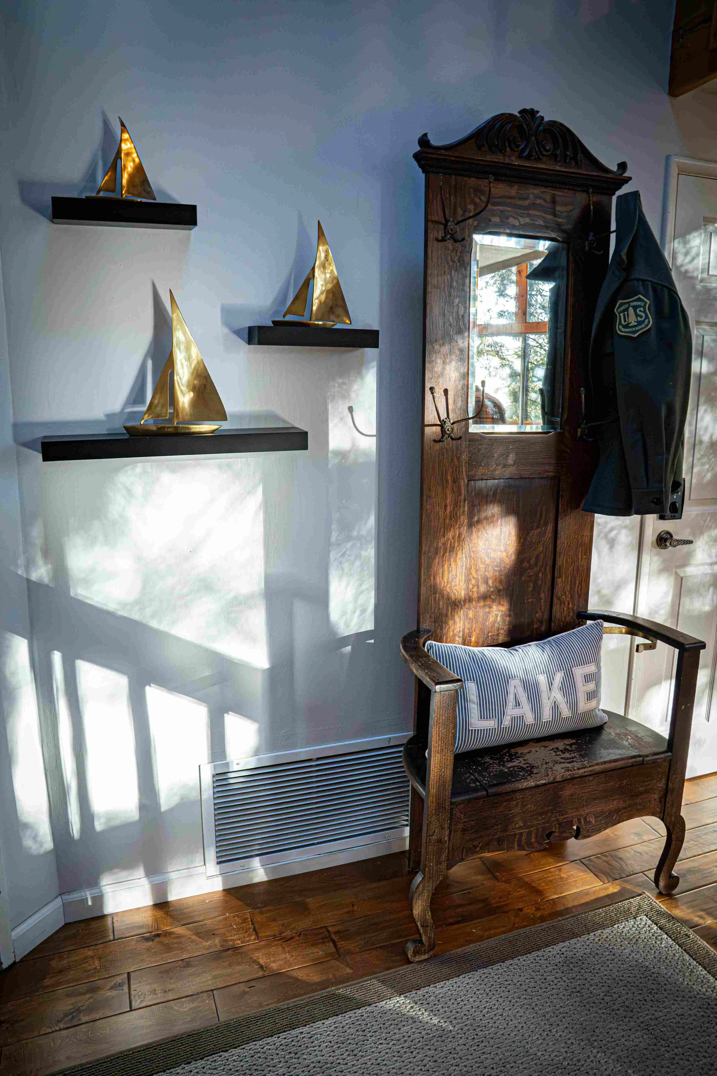 Decorative shelf with three gold sailboat ornaments, small wall hooks on a wooden entryway piece with a mirror and a jacket hanging on it, and a wooden bench with a striped pillow that says 'LAKE'.