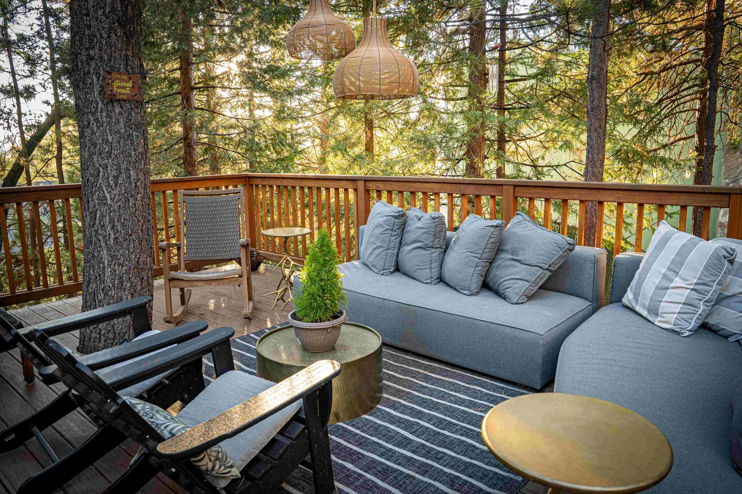 Outdoor balcony with wooden railing surrounded by trees, featuring a gray cushioned sofa with pillows, a small round gold coffee table, a wooden rocking chair with a woven seat, a black wooden chair, a potted plant on a textured round table, and two 