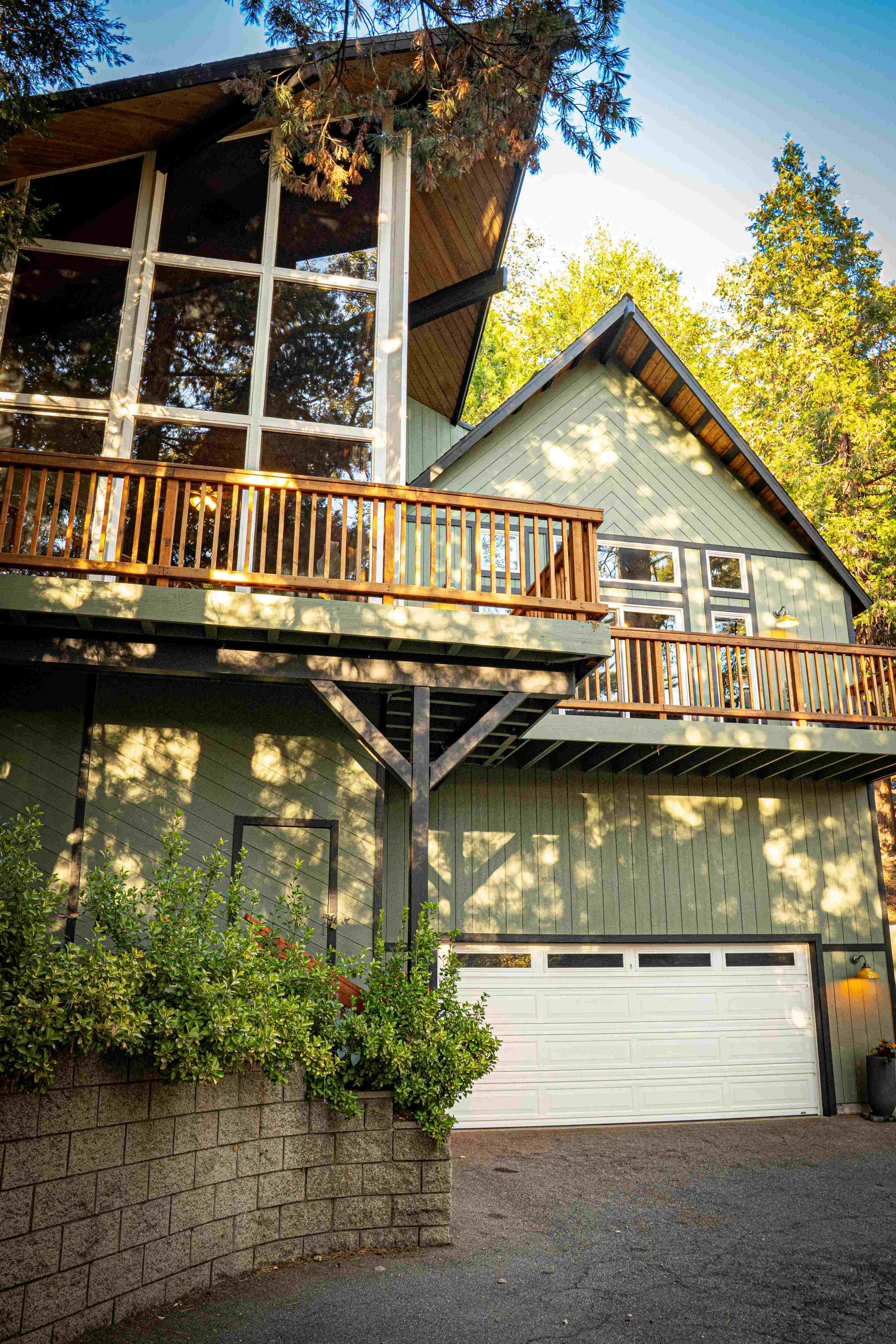 A modern multi-story house with green exterior, large glass windows, a wooden balcony, and a white garage door, surrounded by trees and shrubs.