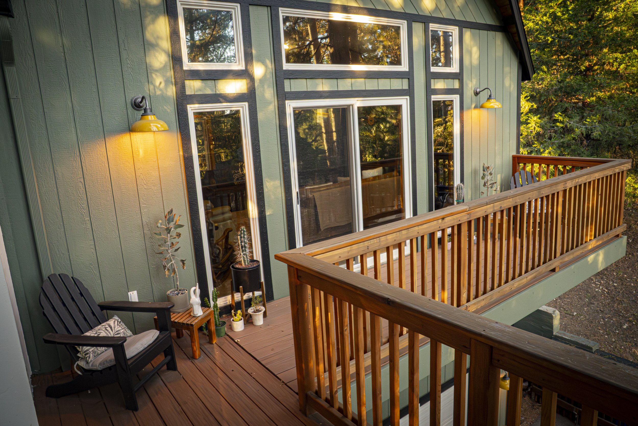 Second story wooden deck with green house siding and large glass windows, decorated with potted plants and yellow wall lights, surrounded by trees.