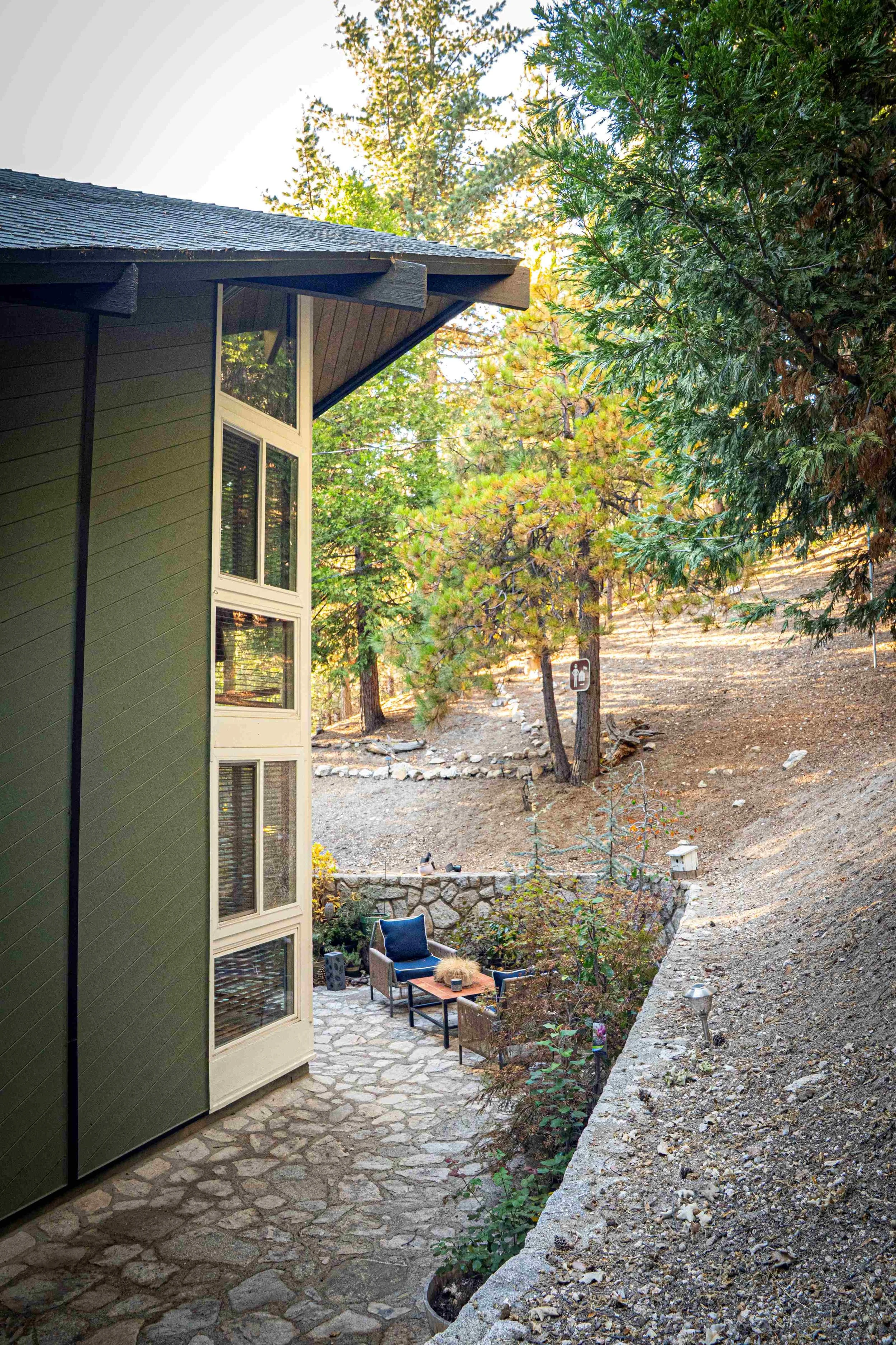 An outdoor patio area beside a house with large windows, surrounded by trees and natural landscape. The patio has a small seating area with two chairs, a small table, and decorative plants.