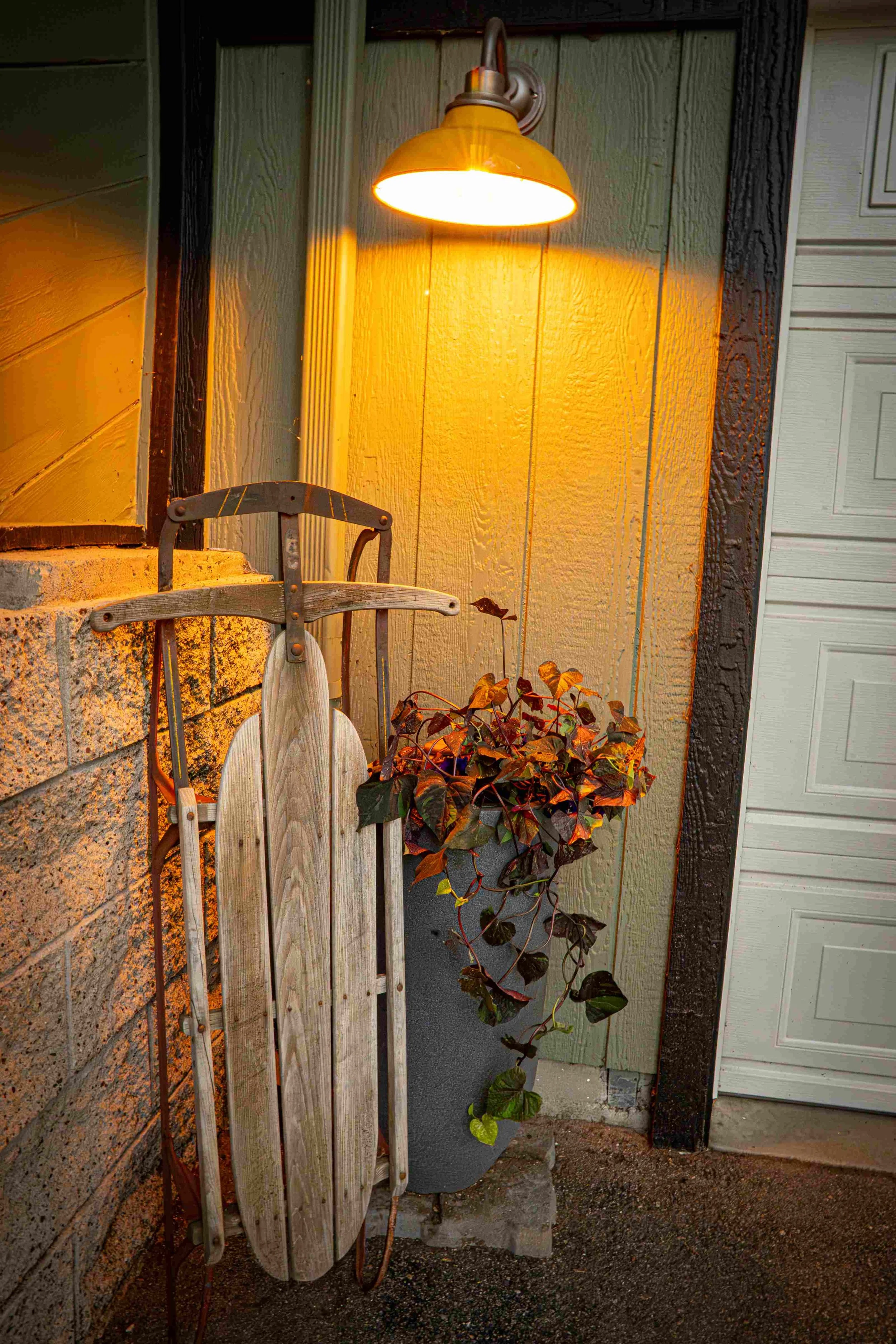 A yellow outdoor wall light illuminating a small patio area with a wooden sled leaning against a brick wall and a potted plant on the ground.