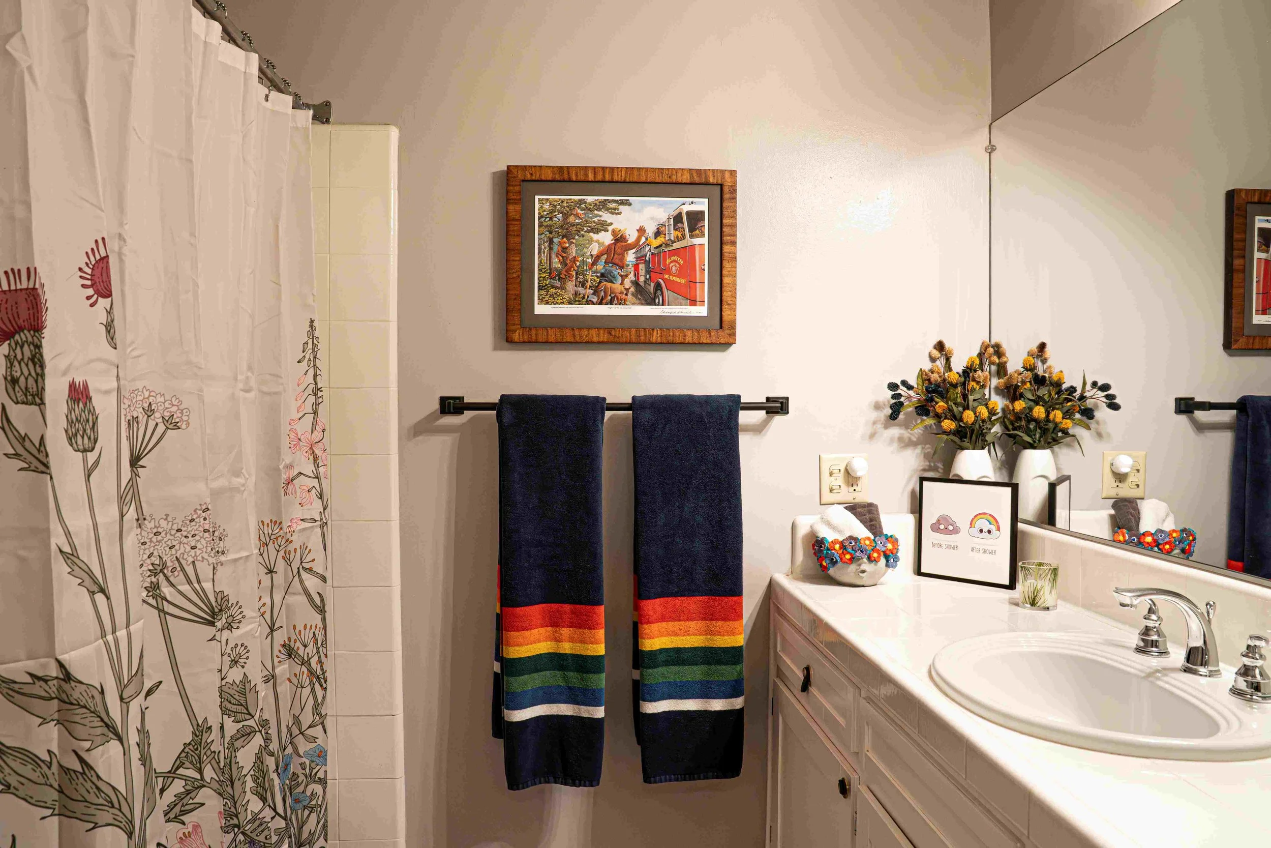 Bathroom with a floral shower curtain, navy towels with rainbow stripes, a framed picture of children at a fire station, a sink with a large mirror, a vase of flowers, and framed artwork with rainbows and clouds.