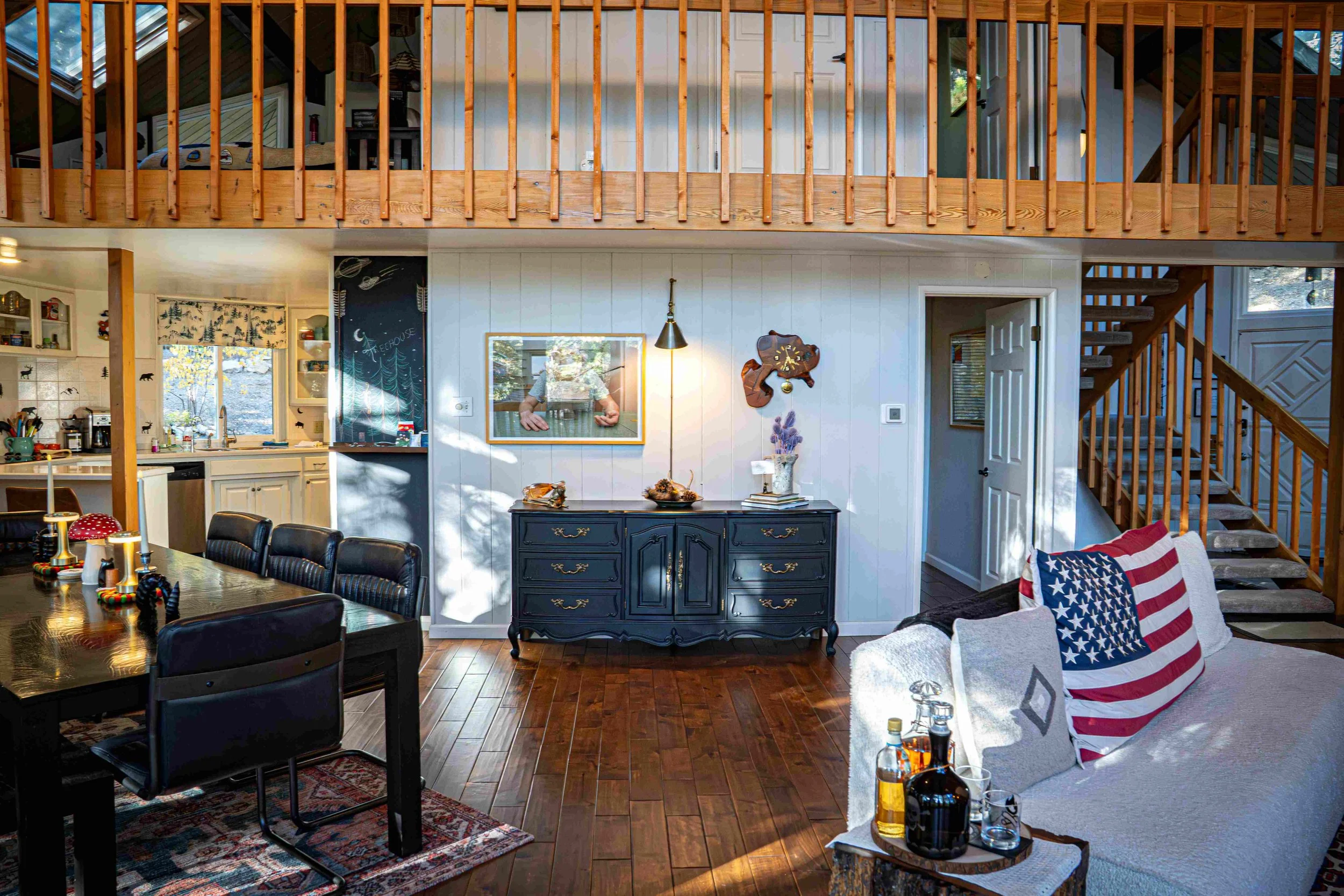 Interior view of a living room with a dining area, featuring a wooden floor, a black sideboard with decorative items, a white sofa with an American flag pillow, and a staircase leading upstairs. The space has a loft area with a railing and a staircas