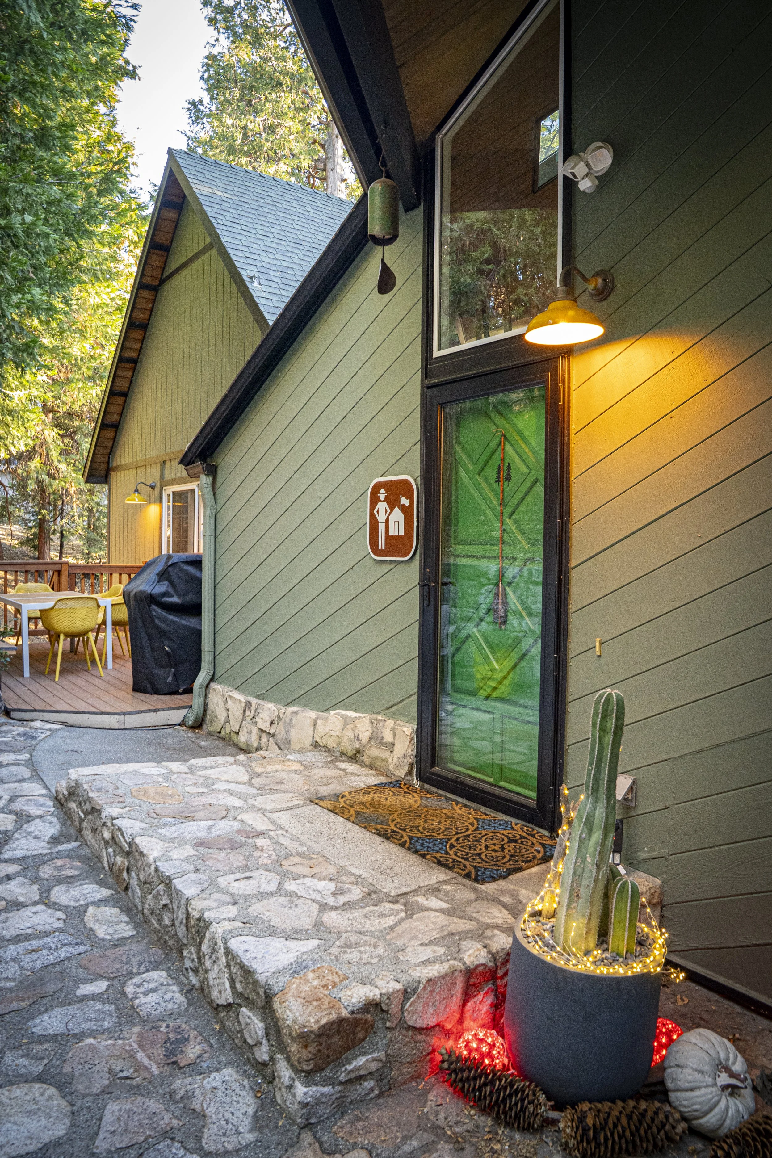Exterior view of a house with a stone pathway leading to a glass door with a decorative green design. Near the door, a potted cactus decorated with string lights and surrounded by pinecones and pumpkins. The house has green siding, outdoor lighting, 