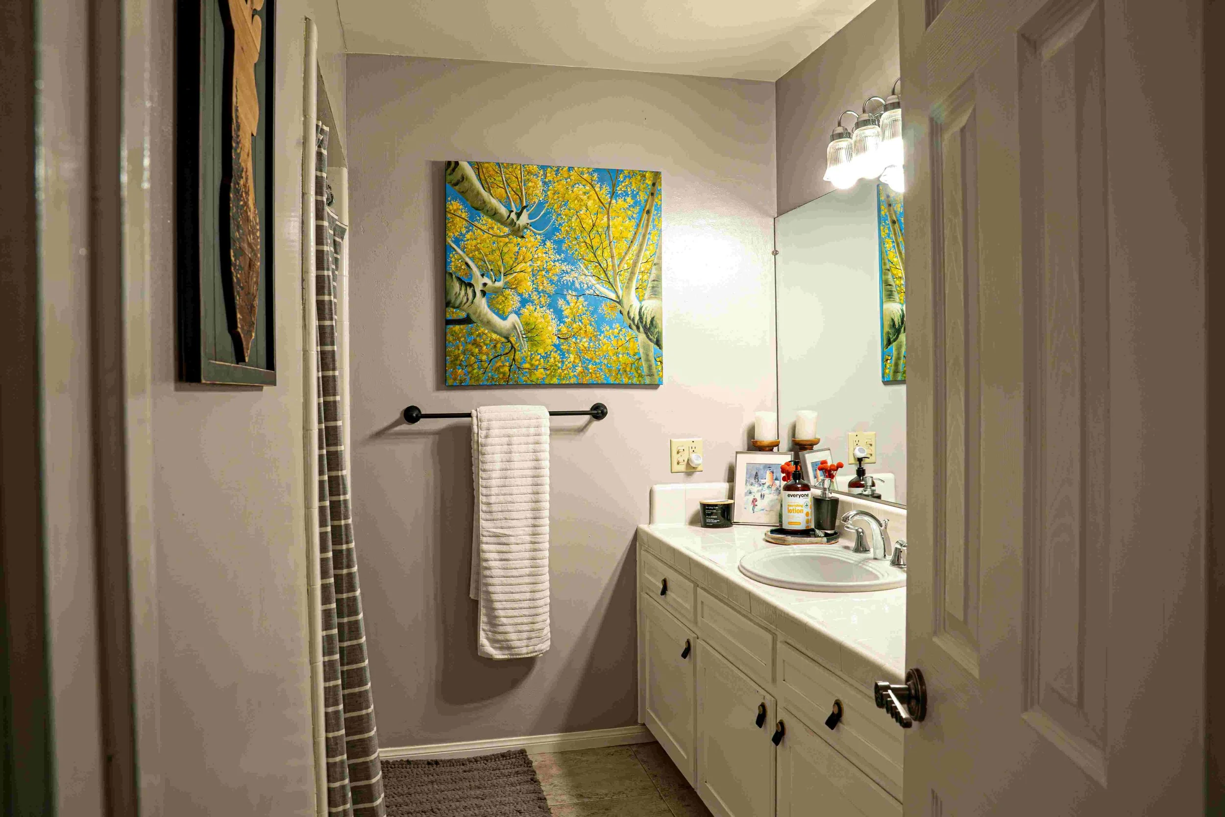 A small bathroom with a white vanity, mirror, and art on the wall above the towel rack. The countertop has toiletries and framed photos, with a light fixture above the mirror. There are candles and a bottle of lotion on the vanity.